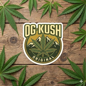 May include: A sticker with the text "OG KUSH ORIGINAL" and a cannabis leaf design, set against a mountain backdrop. The sticker is surrounded by green cannabis leaves, all on a wooden background.