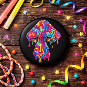 Könnte beinhalten: Ein schwarzer Knopf mit einem psychedelischen Pilzdesign in leuchtenden Farben. Der Pilz hat einen tropfenden Effekt mit Rot-, Gelb-, Blau- und Grüntönen. Der Knopf ist von Partydekorationen auf einer Holzoberfläche umgeben.