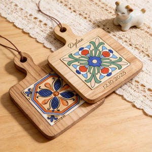 Può includere: Due taglieri in legno con inserti decorativi in piastrelle di ceramica. Una piastrella presenta un disegno floreale blu, l'altra ha un motivo floreale con accenti rossi, blu e verdi. Ogni tagliere ha un manico e un anello in pelle.