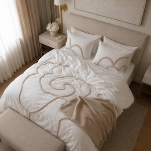 May include: A white bedding set featuring a duvet cover and two pillowcases with raised, beige tufted swirl patterns. A textured beige throw blanket is draped over the foot of the bed, which is positioned against a neutral upholstered headboard.