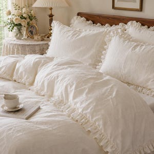 Può includere: Un set di biancheria da letto color crema composto da un copripiumino e due federe abbinate, tutti decorati con delicati bordi a volant. Il tessuto ha un aspetto sottile e testurizzato. Una piccola tazza bianca poggia sopra un libro.