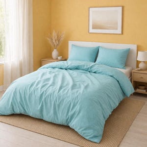 Puede incluir: Una cama con una funda nórdica azul claro y dos fundas de almohada a juego. La cama está sobre una alfombra beige. La habitación tiene paredes amarillas, un cabecero blanco y un cuadro enmarcado.