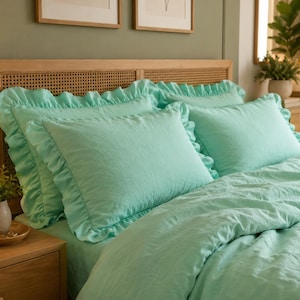 Può includere: Un set di biancheria da letto color verde menta composto da due cuscini rettangolari con bordi arricciati e un copripiumino abbinato. Il tessuto ha un aspetto testurizzato e i cuscini sono posizionati contro una testiera in legno chiaro.