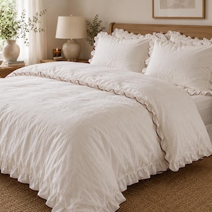 May include: A white duvet cover and two matching pillow shams featuring a textured fabric surface. The bedding set is accented with a delicate ruffled trim along the edges, which is highlighted by a thin, dark brown piping detail throughout.