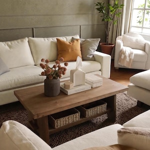 May include: A living room scene featuring a white sofa with orange and gray throw pillows. A wooden coffee table holds a vase of flowers, books, and decorative boxes. Woven baskets are stored on the table's lower shelf. A white armchair and a potted plant are also visible.