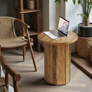 May include: A round wooden side table with a cylindrical base. The table has a natural wood finish and is topped with a laptop and eyeglasses. The table is surrounded by other wooden furniture and plants.