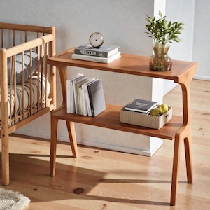 May include: A two-tiered wooden side table with a natural finish. The top shelf holds books, a clock, and a vase with greenery. The lower shelf features books and a woven basket with fruit. The table is next to a wooden crib.