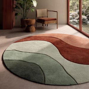 Könnte beinhalten: Ein modernes Wohnzimmer mit einem runden Teppich mit wellenförmigem Design in Grün-, Beige- und Rosttönen. Ein Holzstuhl mit geflochtener Rückenlehne und ein kleiner Holzbeistelltisch sind ebenfalls zu sehen. Eine große Topfpflanze und ein Fenster mit Blick auf eine üppige Landschaft.