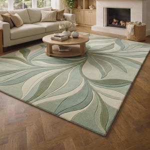 May include: A rectangular area rug featuring a large-scale, flowing leaf pattern in shades of sage green, olive, and cream. The design consists of curved, organic shapes that radiate across the surface, creating a textured, tufted appearance.