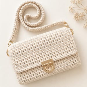 Puede incluir: Un bolso de hombro de ganchillo color crema con cuerpo rectangular y cierre de solapa. El bolso incluye una correa de ganchillo gruesa a juego sujeta con herrajes de metal dorado y un cierre central de metal dorado con detalle de anillo.