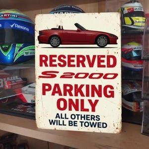 May include: A rectangular metal sign featuring a red convertible car graphic at the top. The text reads RESERVED S2000 PARKING ONLY ALL OTHERS WILL BE TOWED in red and black lettering against a distressed off-white background with rust-like edges.