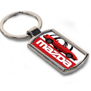 May include: A rectangular silver-toned keychain with a red Mazda sports car design. The keychain features the Mazda logo in white on a red background. The car is red with black tires.