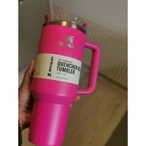 May include: A bright pink Stanley Quencher H2.0 Tumbler with a handle. The tumbler has a clear lid and a silver rim. The label reads "The Flowstate Quencher H2.0 Tumbler 40 oz / 1.18 L All Day Hydration". The Stanley logo is visible.