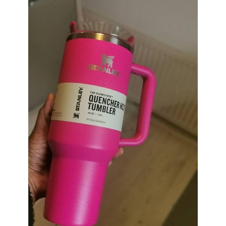 May include: A bright pink Stanley Quencher H2.0 Tumbler with a handle. The tumbler has a clear lid and a silver rim. The label reads "The Flowstate Quencher H2.0 Tumbler 40 oz / 1.18 L All Day Hydration". The Stanley logo is visible.