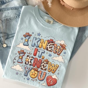 May include: A light blue t-shirt featuring the text I KNEW IT, I KNEW YOU and TS inside a heart. The graphic includes cowboy hats, stars, clouds, and various patterns like cow print and plaid. The shirt is paired with light blue denim jeans and a tan hat.