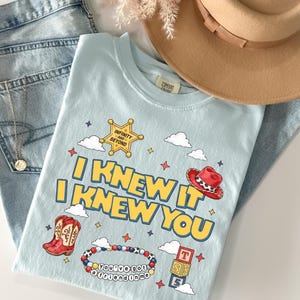 May include: A light blue t-shirt featuring the yellow text I KNEW IT I KNEW YOU, a gold sheriff star with the text INFINITY AND BEYOND, a red cowboy hat, red cowboy boots, a beaded bracelet with the text YOU'VE GOT A FRIEND IN ME, and blocks.