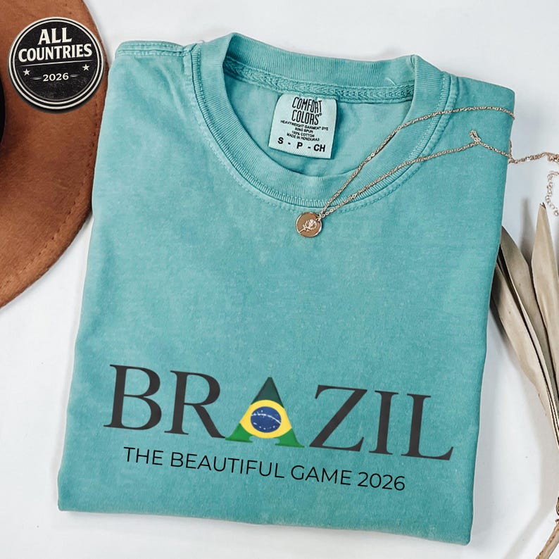 May include: A turquoise t-shirt featuring the word "BRAZIL" in black, with a Brazilian flag design. The text "THE BEAUTIFUL GAME 2026" is below. A gold necklace and a brown hat are also visible in the image.
