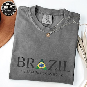May include: A grey t-shirt featuring the word "BRAZIL" in black, with a green and yellow triangle representing the Brazilian flag. The text "THE BEAUTIFUL GAME 2026" is below. A silver necklace with a pendant is on the shirt.