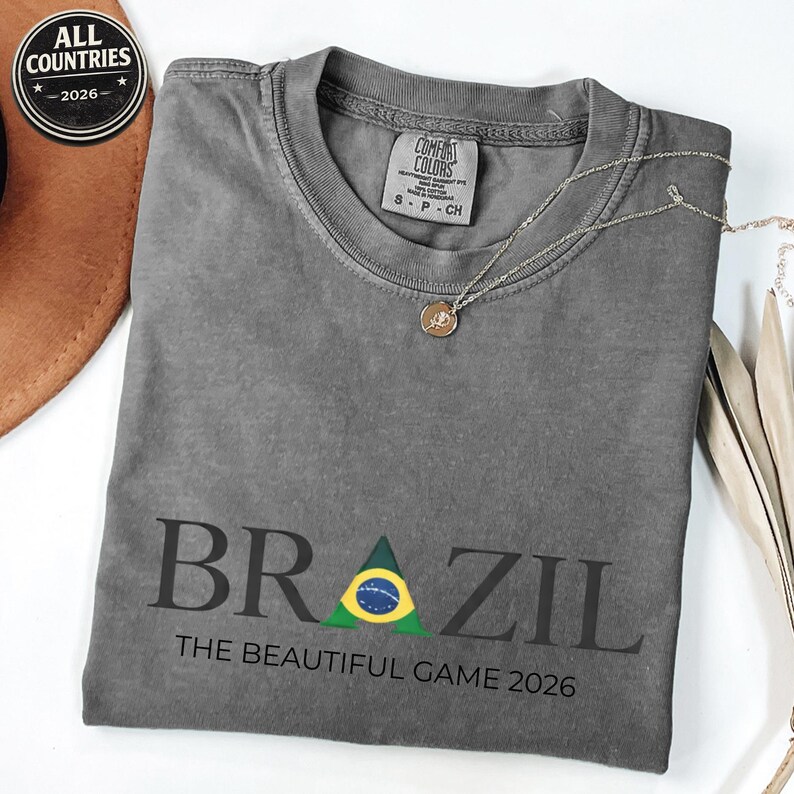 May include: A grey t-shirt featuring the word "BRAZIL" in black, with a green and yellow triangle representing the Brazilian flag. The text "THE BEAUTIFUL GAME 2026" is below. A silver necklace with a pendant is on the shirt.