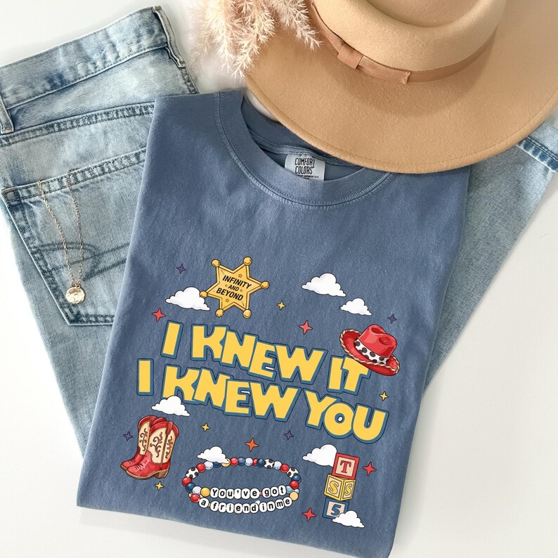 May include: A blue crew neck t-shirt featuring a graphic print with the text I KNEW IT I KNEW YOU, a yellow sheriff star badge with the text INFINITY AND BEYOND, a red cowboy hat, red cowboy boots, a beaded bracelet, and three stacked blocks.