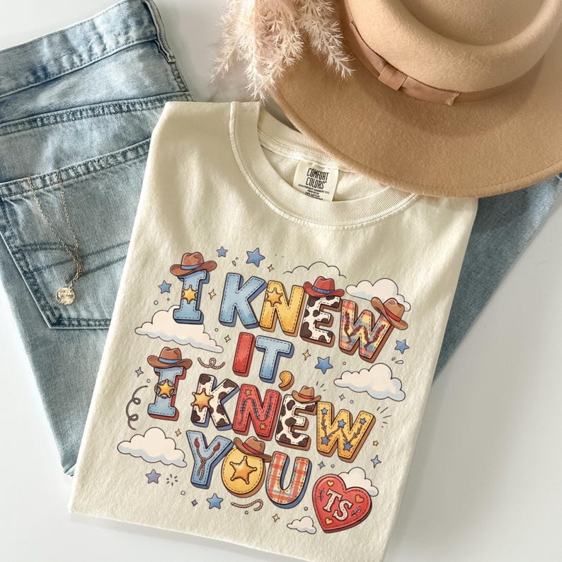 May include: A cream-colored t-shirt featuring the text I KNEW IT, I KNEW YOU with western-themed graphics including cowboy hats, stars, cow print, and a heart with the letters TS, accompanied by light blue denim jeans and a tan felt hat.