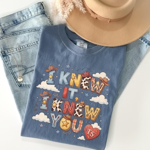May include: A blue t-shirt featuring a graphic print with the text I KNEW IT, I KNEW YOU and TS inside a heart. The design includes cowboy hats, stars, clouds, and cow print patterns. The shirt is paired with light blue denim jeans and a tan hat.