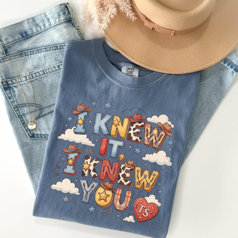 May include: A blue t-shirt featuring a graphic print with the text I KNEW IT, I KNEW YOU and TS inside a heart. The design includes cowboy hats, stars, clouds, and cow print patterns. The shirt is paired with light blue denim jeans and a tan hat.