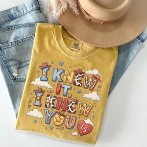 May include: A mustard yellow t-shirt featuring the text I KNEW IT, I KNEW YOU, TS in a western-inspired font with cowboy hat, star, cow print, and plaid patterns. The shirt is displayed with light blue denim jeans, a tan hat, and a silver necklace.