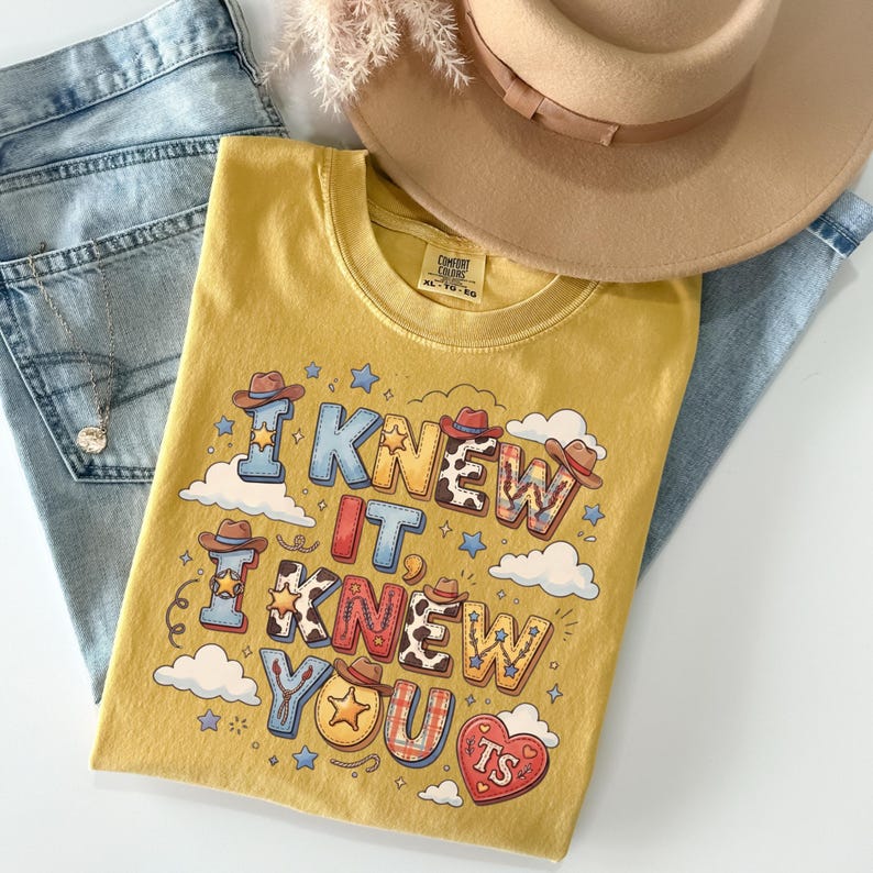 May include: A mustard yellow t-shirt featuring the text I KNEW IT, I KNEW YOU, TS in a western-inspired font with cowboy hat, star, cow print, and plaid patterns. The shirt is displayed with light blue denim jeans, a tan hat, and a silver necklace.
