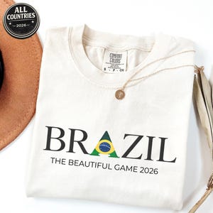 May include: Cream-coloured t-shirt with the word "BRAZIL" in black, featuring a green and yellow graphic. Below, the text reads "THE BEAUTIFUL GAME 2026." A gold necklace and a brown hat are also visible.
