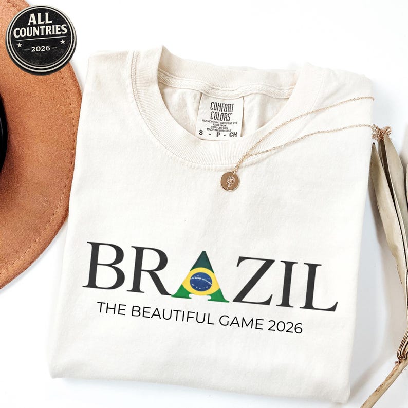 May include: Cream-coloured t-shirt with the word "BRAZIL" in black, featuring a green and yellow graphic. Below, the text reads "THE BEAUTIFUL GAME 2026." A gold necklace and a brown hat are also visible.