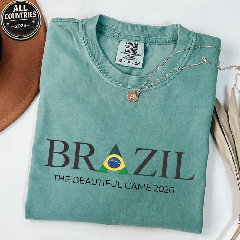 May include: A teal Comfort Colors t-shirt with the word "BRAZIL" in dark letters and a Brazilian flag graphic. Below the word is the text "THE BEAUTIFUL GAME 2026". A silver necklace with a pendant is on the shirt.
