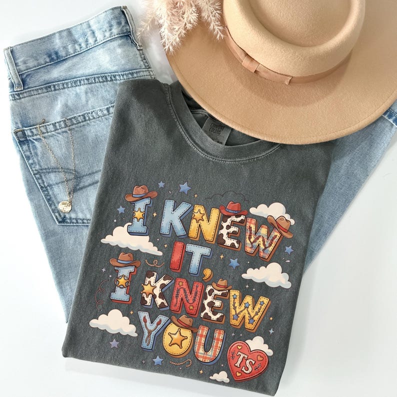 May include: A charcoal gray t-shirt featuring the text I KNEW IT, I KNEW YOU TS in colorful, patterned block letters decorated with cowboy hats, stars, and clouds. The shirt is paired with light blue denim jeans and a tan wide-brimmed hat.