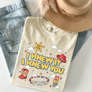 May include: A cream-colored t-shirt featuring a graphic print with the text "I KNEW IT I KNEW YOU" in yellow and blue. The design includes a sheriff badge with "INFINITY AND BEYOND", a red cowboy hat, red boots, a beaded bracelet, and alphabet blocks.