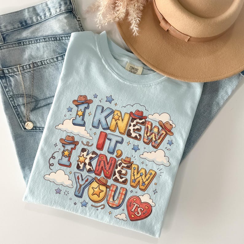 May include: A light blue t-shirt featuring the text I KNEW IT, I KNEW YOU and TS inside a heart. The graphic includes cowboy hats, stars, clouds, and various patterns like cow print and plaid. The shirt is paired with light blue denim jeans and a tan hat.