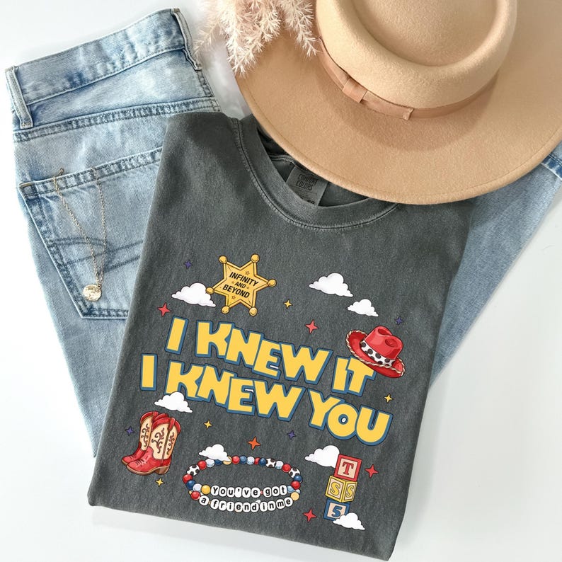 May include: A charcoal gray t-shirt featuring a graphic print with the text I KNEW IT I KNEW YOU, a yellow sheriff star with the text INFINITY AND BEYOND, a red cowboy hat, red cowboy boots, a beaded bracelet, and stacked blocks with the text YOU'VE GOT A FRIEND IN ME.