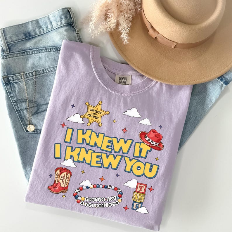 May include: A light purple t-shirt featuring a graphic print with the text I KNEW IT I KNEW YOU, a yellow sheriff star with the text INFINITY AND BEYOND, a red cowboy hat, red cowboy boots, a beaded bracelet, and alphabet blocks, paired with blue jeans and a tan hat.