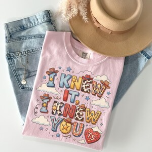 May include: A light pink t-shirt featuring the text I KNEW IT, I KNEW YOU TS in colorful, patterned block letters decorated with small cowboy hats, stars, and clouds. The shirt is displayed alongside a tan wide-brim hat and blue denim jeans.