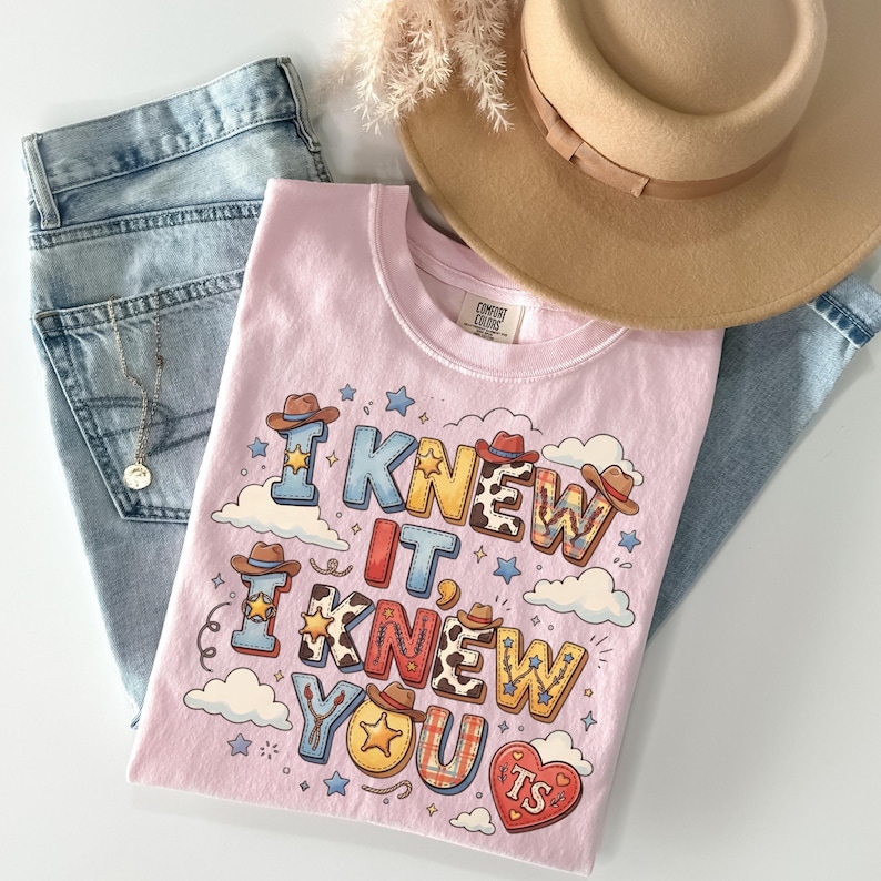 May include: A light pink t-shirt featuring the text I KNEW IT, I KNEW YOU TS in colorful, patterned block letters decorated with small cowboy hats, stars, and clouds. The shirt is displayed alongside a tan wide-brim hat and blue denim jeans.