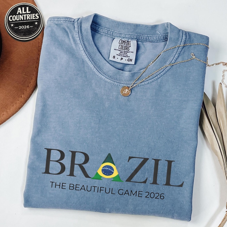 May include: A blue t-shirt featuring the word "BRAZIL" in black with a Brazilian flag design. The text "THE BEAUTIFUL GAME 2026" is below. A gold necklace with a pendant rests on the shirt. The shirt has a Comfort Colors label.