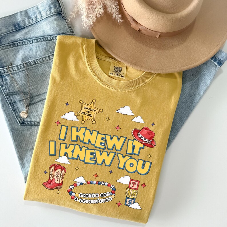May include: A mustard yellow t-shirt featuring the text I KNEW IT I KNEW YOU in blue letters. The graphic includes a sheriff star with the text INFINITY AND BEYOND, a red cowboy hat, red cowboy boots, a beaded bracelet, and stacked alphabet blocks.