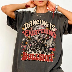 Könnte beinhalten: Ein anthrazitfarbenes Kurzarm-T-Shirt mit einem Grafikdruck, der den Text Dancing Is Everything und Everything Else Is Bullshit zeigt. Das Design umfasst eine Discokugel, tanzende Figuren, Schallplatten, einen Cocktail und Sterne.