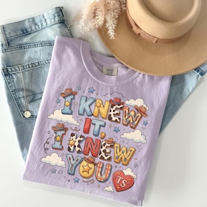 May include: A light purple t-shirt featuring the text I KNEW IT, I KNEW YOU TS in colorful, patterned block letters decorated with cowboy hats, stars, and clouds. The shirt is paired with light blue denim jeans, a tan wide-brimmed hat, and a necklace.