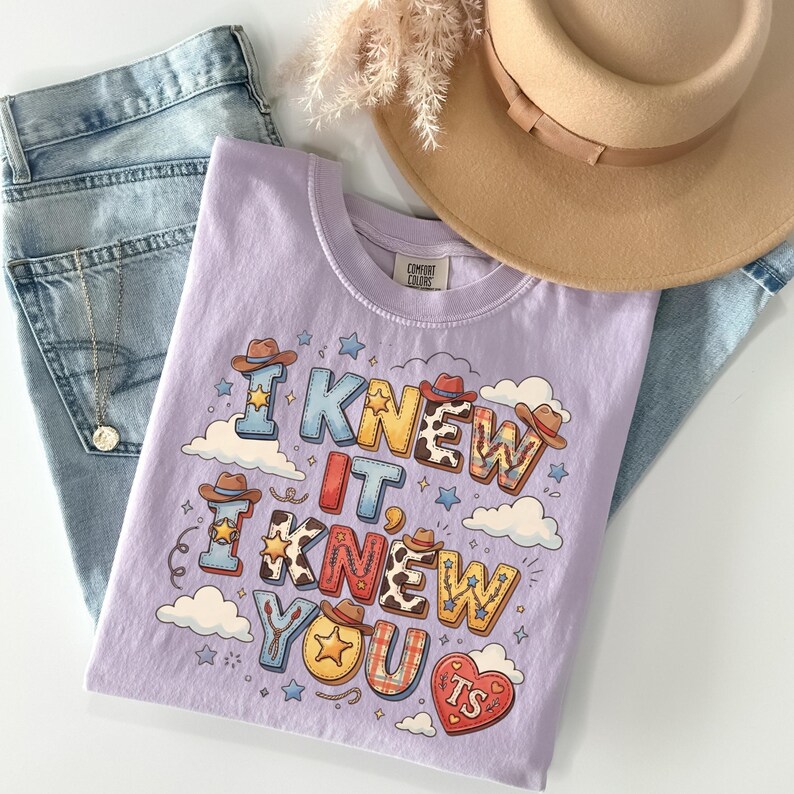 May include: A light purple t-shirt featuring the text I KNEW IT, I KNEW YOU TS in colorful, patterned block letters decorated with cowboy hats, stars, and clouds. The shirt is paired with light blue denim jeans, a tan wide-brimmed hat, and a necklace.