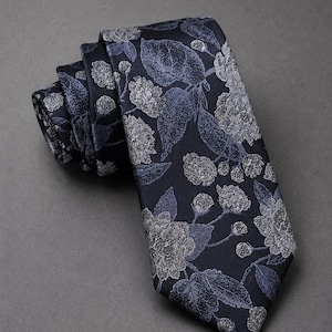 Può includere: Una cravatta blu navy scuro con un motivo floreale ripetuto in tonalità grigio chiaro e blu tenue. Il design presenta illustrazioni dettagliate di fiori in fiore, boccioli e foglie sull'intera superficie del tessuto.