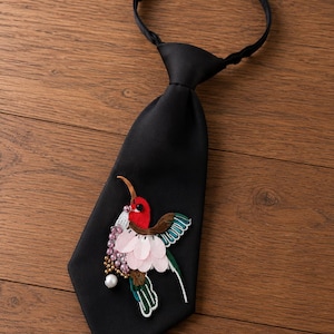 Peut inclure: Une cravate noire ornée d'une broderie représentant un colibri. L'oiseau est réalisé avec des fils rouges, verts et marron, agrémenté de sequins rose pâle en forme de pétales et de petites perles rondes irisées formant un amas près du poitrail.