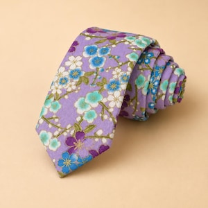 May include: A necktie in a light purple fabric featuring a repeating floral pattern of white, turquoise, and dark purple blossoms with green stems. The tie is rolled into a spiral shape, displaying the pointed end and the layered fabric folds.