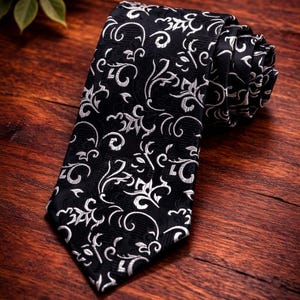 Può includere: Cravatta nera con motivo floreale bianco. La cravatta è arrotolata e presentata su una superficie di legno. Il design presenta elementi floreali a vortice, creando un look classico ed elegante.