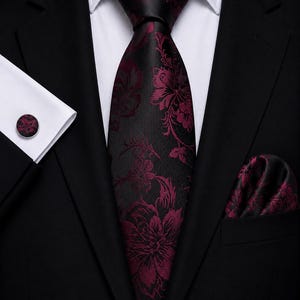 Op de afbeelding: Een zwarte das met een donker magenta bloemenpatroon, een bijpassende pochet en een enkele ronde manchetknop. De set wordt gedragen met een wit overhemd en een zwart colbert, wat zorgt voor een formeel ensemble met een donker kleurenpalet.