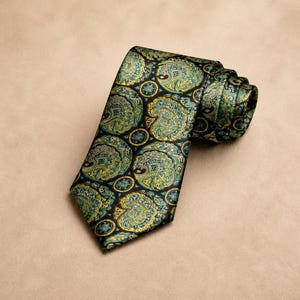 Può includere: Una cravatta arrotolata con un motivo paisley nei toni del verde, verde acqua e oro su sfondo nero. La cravatta è realizzata in un tessuto lucido e viene esposta su una superficie di colore neutro.